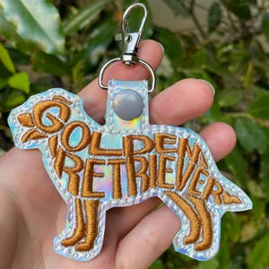 May include: A Golden Retriever keychain with the breed name embroidered in gold thread. The keychain has a silver clasp and a holographic background. The dog shape is outlined in white thread.