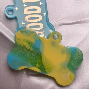 May include: A dog bone-shaped pet tag with a yellow, green, and blue swirled design. The tag has a hole for attaching to a collar. A light blue bone-shaped tag with the words "GOOD BOY" is in the background.