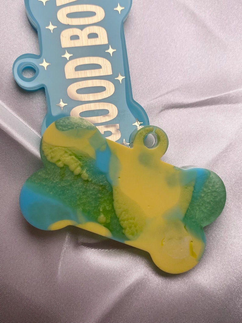 May include: A dog bone-shaped pet tag with a yellow, green, and blue swirled design. The tag has a hole for attaching to a collar. A light blue bone-shaped tag with the words "GOOD BOY" is in the background.