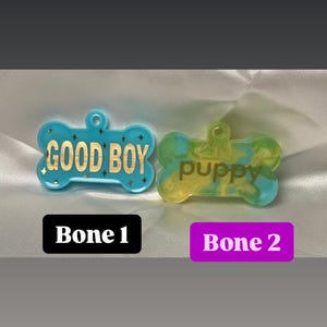 May include: Two bone-shaped pet tags. One is light blue with gold text reading "GOOD BOY" and star accents. The other is yellow and blue with "puppy" in a script font. The tags are on a white satin surface.
