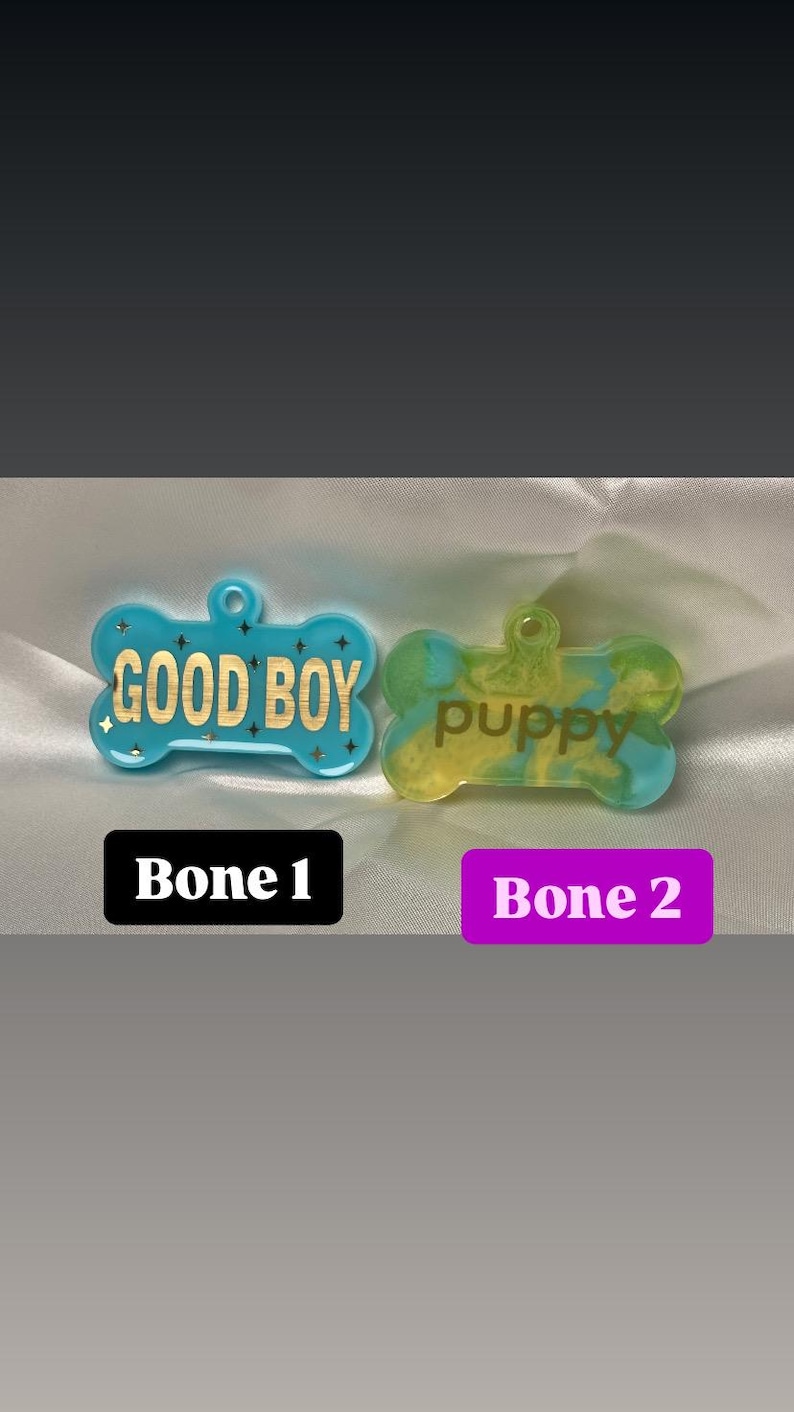 May include: Two bone-shaped pet tags. One is light blue with gold text reading "GOOD BOY" and star accents. The other is yellow and blue with "puppy" in a script font. The tags are on a white satin surface.