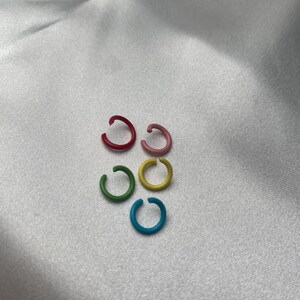 May include: Six colorful ear cuffs in red, pink, green, yellow, and blue are arranged on a white, silky surface. The ear cuffs are small, circular, and appear to be made of metal or a similar material. The colors are vibrant and eye-catching.