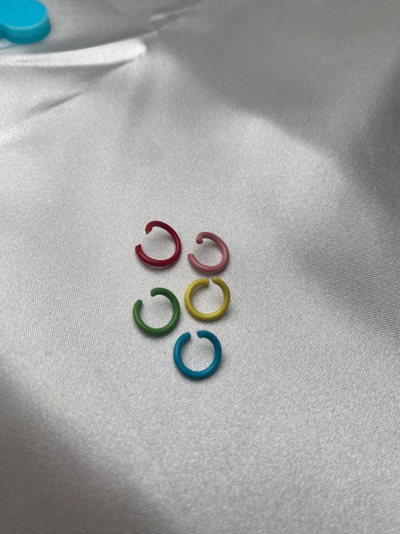 May include: Six colorful ear cuffs in red, pink, green, yellow, and blue are arranged on a white, silky surface. The ear cuffs are small, circular, and appear to be made of metal or a similar material. The colors are vibrant and eye-catching.