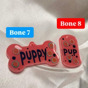 May include: Two pink pet tags with a watermelon design. One tag is bone-shaped and says "PUPPY" in dark blue letters. The other tag is rectangular and says "PUP." Both tags have a hole for attaching to a collar.