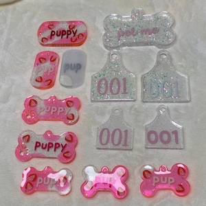 May include: A selection of pet tags in different shapes and colours. Some tags are rectangular, bone-shaped, or tag-shaped. The tags are pink, clear, and white with glitter and the words "puppy", "pup", "pet me", and the number "001".