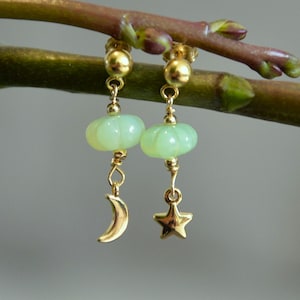 Puede incluir: Un par de pendientes dorados con temática celestial. Cada pendiente presenta una cuenta verde claro en forma de calabaza, un dije de media luna dorada y un dije de estrella dorada. Los pendientes cuelgan de un perno de bola dorada.