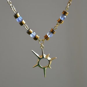 Puede incluir: Un collar de oro con un colgante de estrella y cuentas azules. El collar está hecho de una cadena de oro con pequeñas cuentas azules redondas.