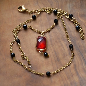 Peut inclure: Un collier en chaîne doré avec un pendentif rouge rectangulaire facetté. Le collier comporte des perles noires réparties le long de la chaîne. Le pendentif est encadré d'or et une petite perle noire pend en dessous. Un fermoir sécurise le collier.