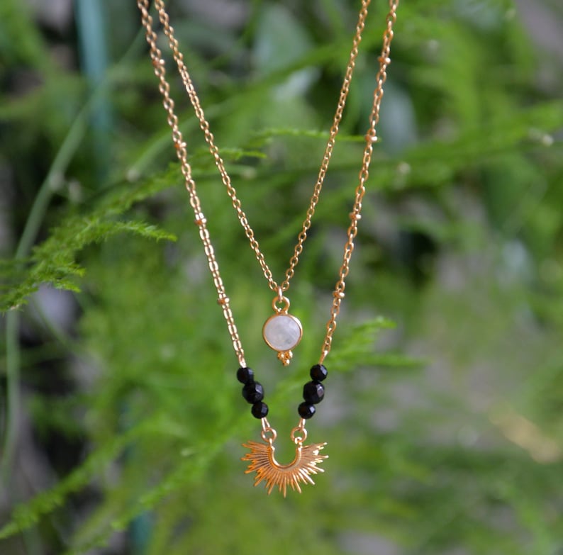 May include: A gold layered necklace with a moonstone pendant and black beads. The bottom layer features a sunburst design.