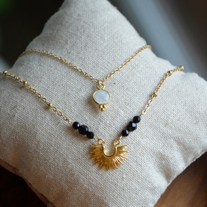 May include: Two gold necklaces with a moonstone pendant and a sunburst pendant. The moonstone pendant is on a delicate chain with small beads. The sunburst pendant is on a chain with black beads.