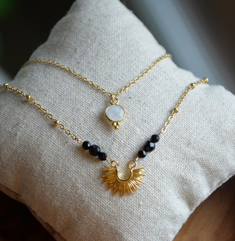 May include: Two gold necklaces with a moonstone pendant and a sunburst pendant. The moonstone pendant is on a delicate chain with small beads. The sunburst pendant is on a chain with black beads.