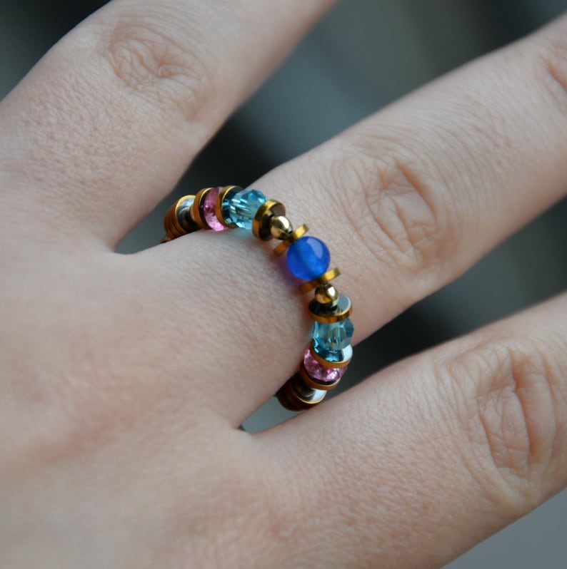 May include: A beaded ring with a gold-coloured band and a pattern of pink, blue, and clear beads. A central blue bead is also present. The ring is worn on a finger.
