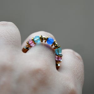 May include: A handmade ring with a bronze band and colourful beads. The ring features pink, light blue, and dark blue beads, interspersed with small gold-coloured accents. The ring is worn on a finger.