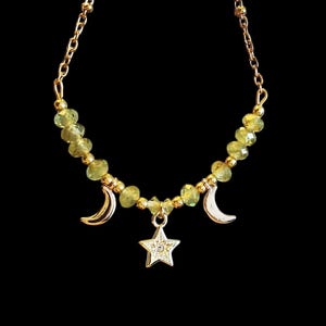May include: A gold necklace with a celestial design. The necklace has a chain with green gemstone beads and gold details. It features three charms: two crescent moons and a star with a clear gem centre. The background is black.