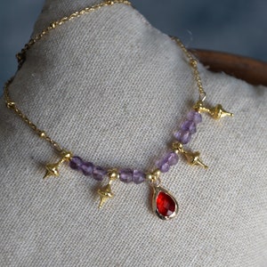 May include: A gold chain necklace with a red teardrop pendant and purple beads. The necklace has four gold charms shaped like small cones.