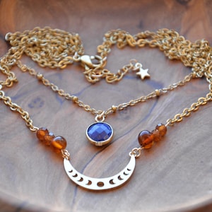Può includere: Una collana a catena dorata con un ciondolo a forma di luna crescente e un ciondolo di pietra preziosa blu. La collana presenta anche perle ambrate.