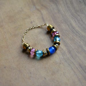May include: A gold chain ring adorned with a selection of colourful beads. The ring incorporates pink, blue, and gold beads, alongside a single blue bead. The ring is displayed on a wooden surface.