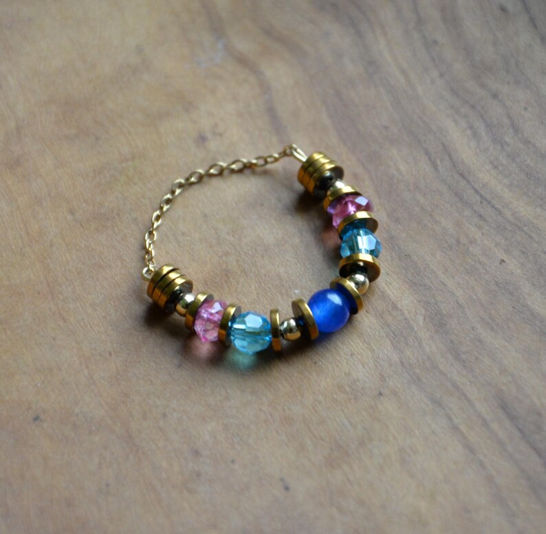 May include: A gold chain ring adorned with a selection of colourful beads. The ring incorporates pink, blue, and gold beads, alongside a single blue bead. The ring is displayed on a wooden surface.