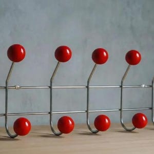 May include: A stylish coat rack featuring six bright red ball hooks on a silver metal frame. The rack is designed for wall mounting and is suitable for hanging coats, hats, or other accessories. The rack's dimensions are approximately 60 cm in length.