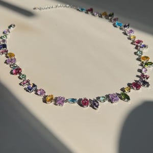 Può includere: Una collana d'argento con una varietà di pietre preziose colorate. Le pietre preziose sono nei toni del rosa, viola, giallo, blu e verde. La collana è su una superficie bianca.
