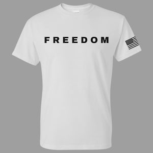 May include: White t-shirt with the word "FREEDOM" printed in large, black letters across the chest. A small American flag is printed on the right sleeve. The shirt has a crew neck and short sleeves.