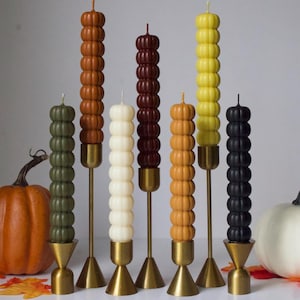 May include: A collection of pillar candles in various colors, including green, orange, white, brown, yellow, and black. Each candle has a pumpkin-like design and is set on a gold-colored metal stand. Pumpkins and fall leaves are also in the image.