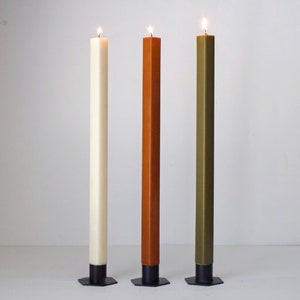 May include: Three tall candles, one white, one orange, and one green, are burning on black metal bases.