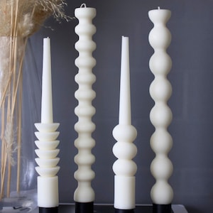 May include: Four white pillar candles with different textured designs. The candles are on black hexagonal bases and are arranged on a book.