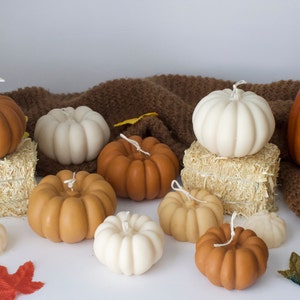 Puede incluir: Una colecci&oacute;n de velas con forma de calabaza de tem&aacute;tica oto&ntilde;al en varios tonos de marr&oacute;n, blanco y naranja. Las velas est&aacute;n dispuestas sobre una superficie de punto marr&oacute;n y balas de heno.