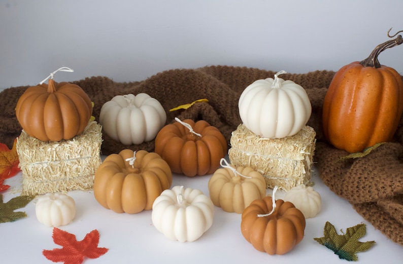 Puede incluir: Una colecci&oacute;n de velas con forma de calabaza de tem&aacute;tica oto&ntilde;al en varios tonos de marr&oacute;n, blanco y naranja. Las velas est&aacute;n dispuestas sobre una superficie de punto marr&oacute;n y balas de heno.