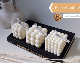 Puede incluir: Un conjunto de tres velas blancas en forma de cubo con un diseño texturizado con forma de burbujas. Las velas están dispuestas sobre una superficie negra y tienen una etiqueta blanca con el texto "MarneyAsh" en cada una. El texto "3 Piece Candle Set" y "100% Natural Soy Wax" se muestra encima de las velas.