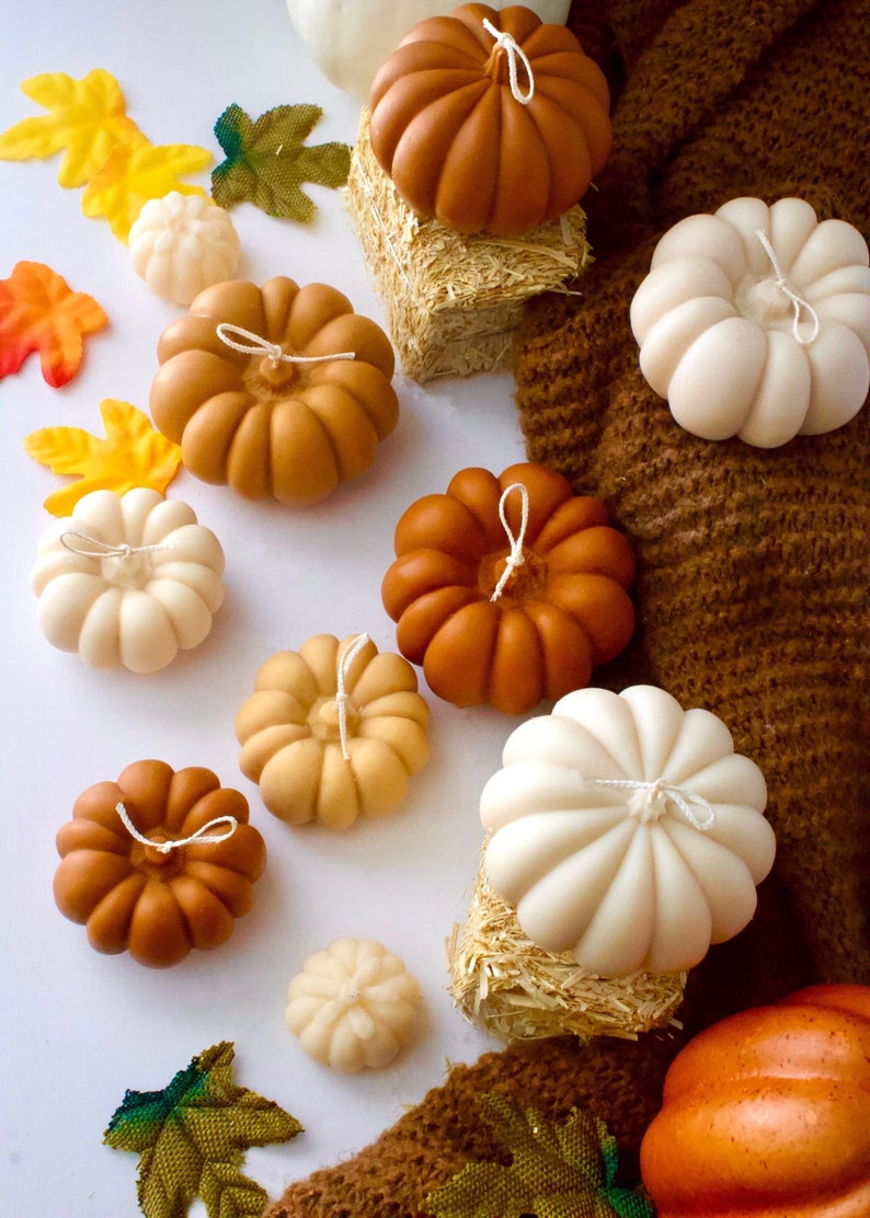 Puede incluir: Una colecci&oacute;n de peque&ntilde;as velas con forma de calabaza hechas a mano en varios tonos de naranja, crema y blanco. Las velas est&aacute;n dispuestas sobre una superficie blanca con decoraciones de oto&ntilde;o.