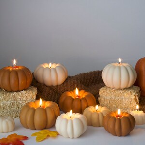 Puede incluir: Un grupo de velas encendidas con forma de calabaza en varios tonos de marr&oacute;n y blanco, dispuestas sobre una manta de punto marr&oacute;n y beige. Las velas est&aacute;n rodeadas de peque&ntilde;as hojas de oto&ntilde;o y una sola calabaza entera.