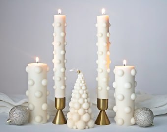 Puede incluir: Cinco velas blancas en forma de pilar con un diseño texturizado con forma de burbujas. Las velas están encendidas y están dispuestas en fila sobre una superficie blanca. Dos de las velas están sobre candelabros de metal dorado. Una vela blanca más pequeña con forma de árbol de Navidad está en el centro.