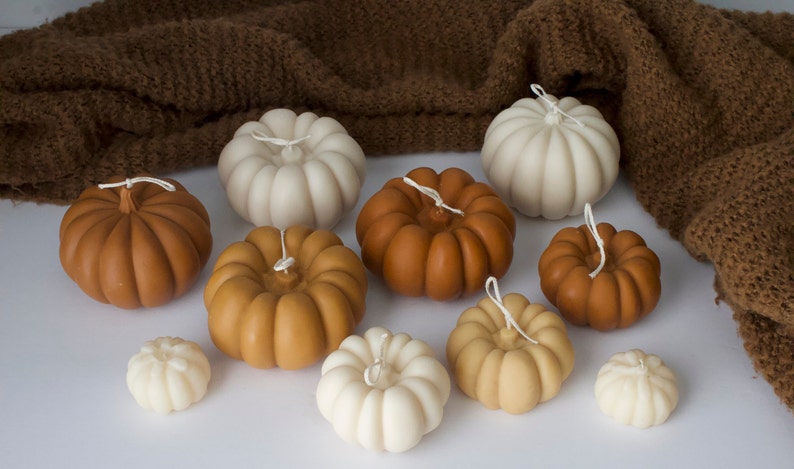 Puede incluir: Una colecci&oacute;n de nueve peque&ntilde;as velas con forma de calabaza en varios tonos de marr&oacute;n, beige y blanco. Las velas est&aacute;n dispuestas sobre una superficie blanca y est&aacute;n rodeadas por una manta de punto marr&oacute;n.