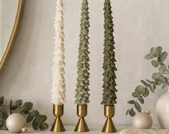 May include: Three tall, slender taper candles shaped like pine trees, each standing in a polished brass candlestick holder. One candle is cream-colored, while the other two are in varying shades of sage and forest green, featuring textured layers.