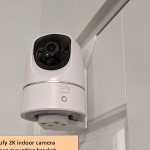 May include: A white Eufy 2K indoor security camera mounted on a door frame. The camera has a black lens and the word "eufy" is printed on the front. The camera is attached to a white door mounting bracket.