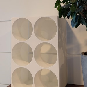 May include: A cream-coloured wine rack with six circular openings. The rack is made of a solid material and sits on a wooden shelf. A potted plant with green leaves is visible in the background.