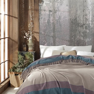 May include: A beige blanket with a blue border and tassels is draped over a bed. The blanket has a woven texture and a stripe of red and purple. The bed has white pillows and a neutral-toned headboard.