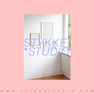May include: A white wall with three framed grid-like designs. The text "STYKKE*STUDIO" is written in blue letters on the wall. The text "NOW" is written in a circle on the floor.