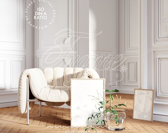 May include: A white armchair with a tufted back and a wooden floor. Two picture frames are on the floor, one is empty and the other has a gold frame. A plant in a glass vase is in front of the empty frame.