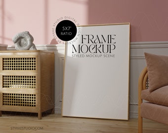 May include: A gold frame mockup with a white background, sitting on a wooden floor in a styled room with a pink wall, a wooden cabinet, and a wicker chair with a pink cushion. The frame is 5x7 inches.