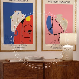 May include: Two framed posters with abstract art designs, one with red, yellow, and blue colors and the text "Prepared Smart Objects", the other with pink, blue, and black colors and the text "Easy to Use Mockup", a white ceramic lamp with a face design, and a stack of magazines on a wooden table.