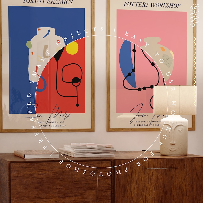 May include: Two framed posters with abstract art designs, one with red, yellow, and blue colors and the text "Prepared Smart Objects", the other with pink, blue, and black colors and the text "Easy to Use Mockup", a white ceramic lamp with a face design, and a stack of magazines on a wooden table.
