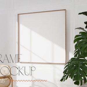 Puede incluir: Un marco cuadrado y en blanco con un borde de madera clara cuelga de una pared blanca. El texto "FRAME MOCKUP" se muestra en una fuente negra elegante y delgada. A la derecha hay una gran planta verde con hojas anchas y a la izquierda una silla de mimbre.