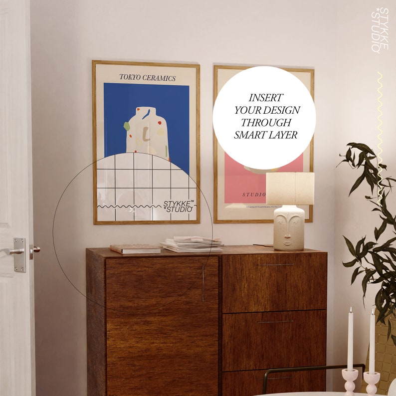 May include: Two framed posters on a white wall above a wooden dresser. The poster on the left has a blue background with a white and red abstract design and the text "TOKYO CERAMICS". The poster on the right has a pink background with the text "INSERT YOUR DESIGN THROUGH SMART LAYER" and "STUDIO".
