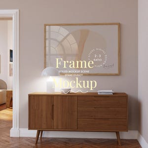 Puede incluir: Una cómoda de madera con tres cajones, una lámpara blanca con una pantalla blanca y una impresión enmarcada con un diseño abstracto beige y marrón. La impresión tiene el texto "Frame Styled Mockup Scene Stykke Studio™ 2:3 Ratio Digital Design Mockup".