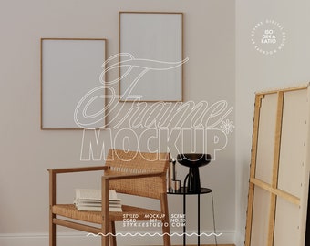 May include: Two framed blank art prints with gold trim hang on a beige wall. A woven chair with a stack of books sits below the frames. A black lamp and side table are next to a large canvas. The image text says "Frame Mockup".