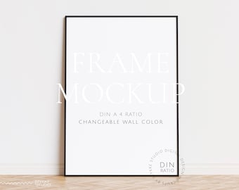 Könnte beinhalten: Ein schwarzer Rahmen-Mockup mit dem Text "FRAME MOCKUP" und "DIN A 4 RATIO CHANGEABLE WALL COLOR" an einer weißen Wand. Der Rahmen steht auf einem Holzboden.