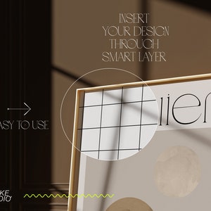 May include: A gold framed poster with a white background and a black grid pattern. The poster has the text "illier" in black and a beige circle with a textured surface.